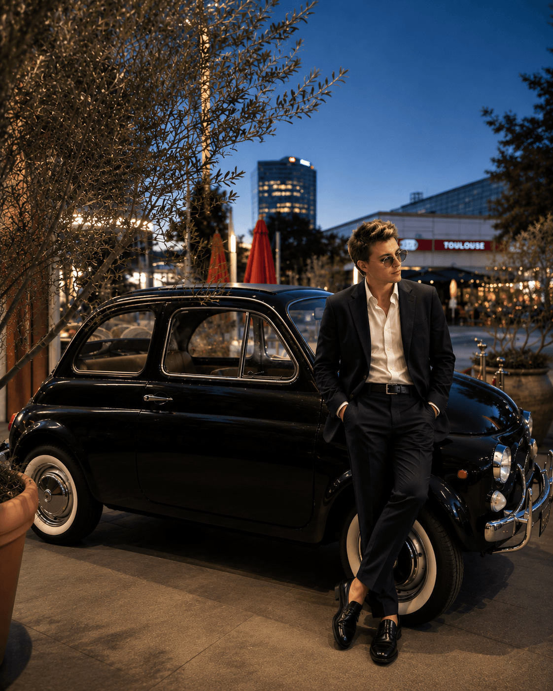 Hombre con traje elegante apoyado en un coche clásico negro bajo las luces nocturnas.